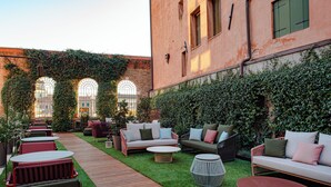 Un patio al aire libre con cubierta de madera, diversas opciones de asientos que incluyen sofás y sillones, y un muro de ladrillo con ventanas arqueadas.