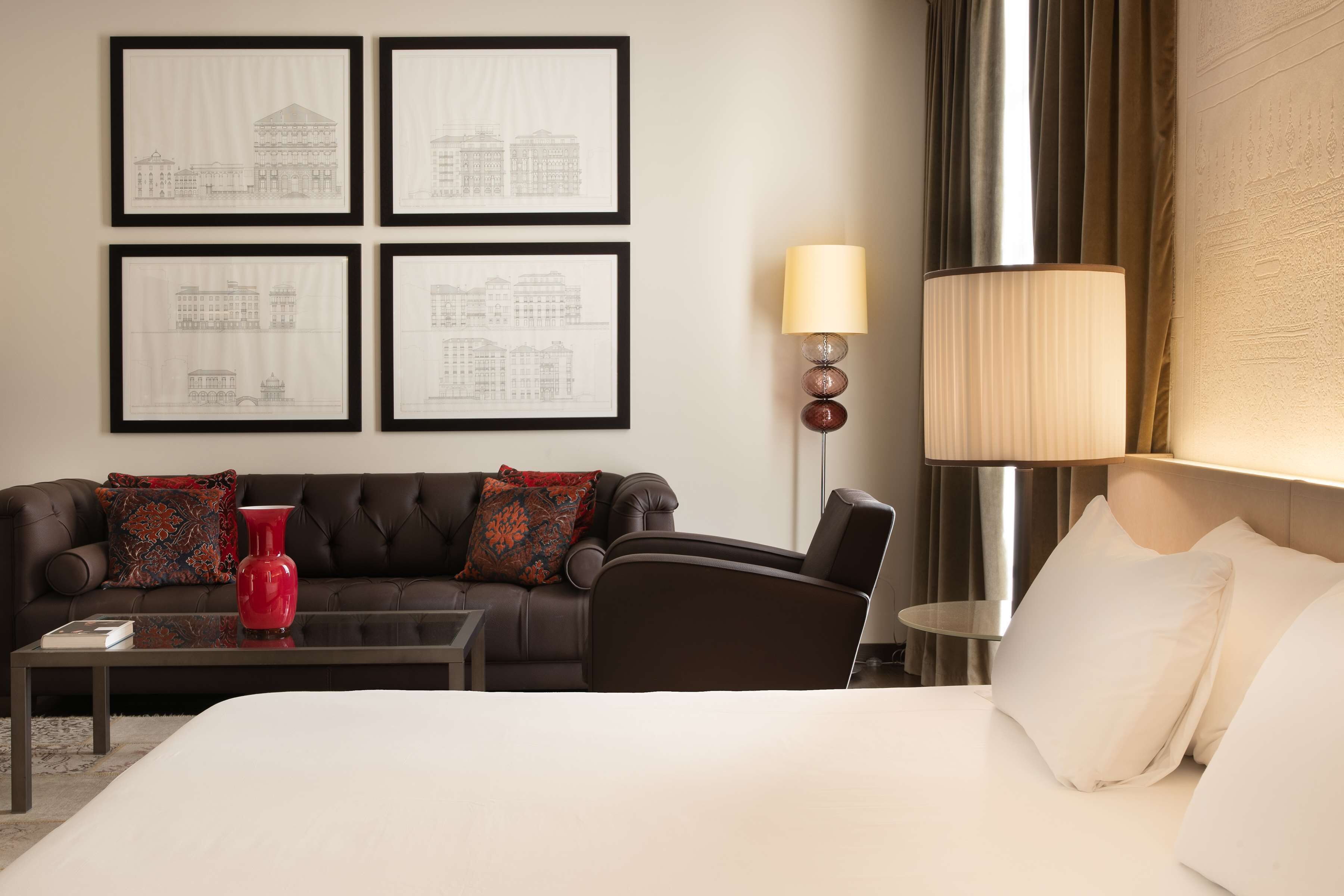 A hotel room with a bed, a sofa, a coffee table, and a lamp. There are four framed architectural drawings on the wall.