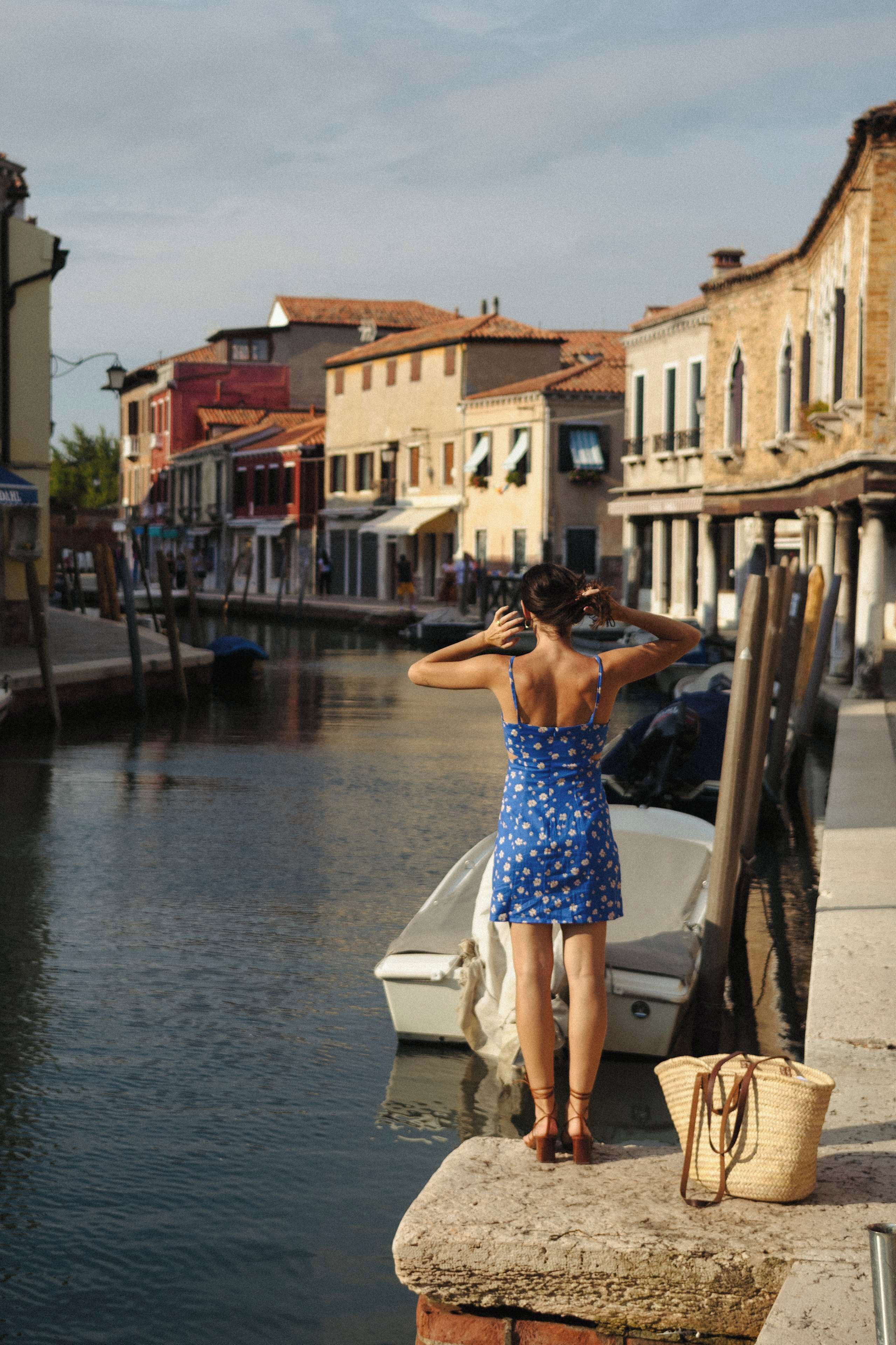 Mulher de vestido azul parada à beira de um canal, com edifícios ao fundo.