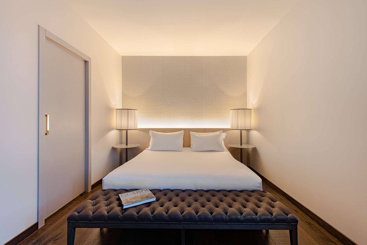 Une chambre d’hôtel bien aménagée, avec un grand lit, deux lampes de chevet, un banc aux tons sombres et une porte.