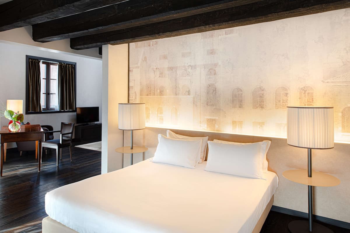 Une chambre d’hôtel avec un grand lit, deux lampes de chevet, un bureau avec une chaise et un mur orné d’une fresque historique.