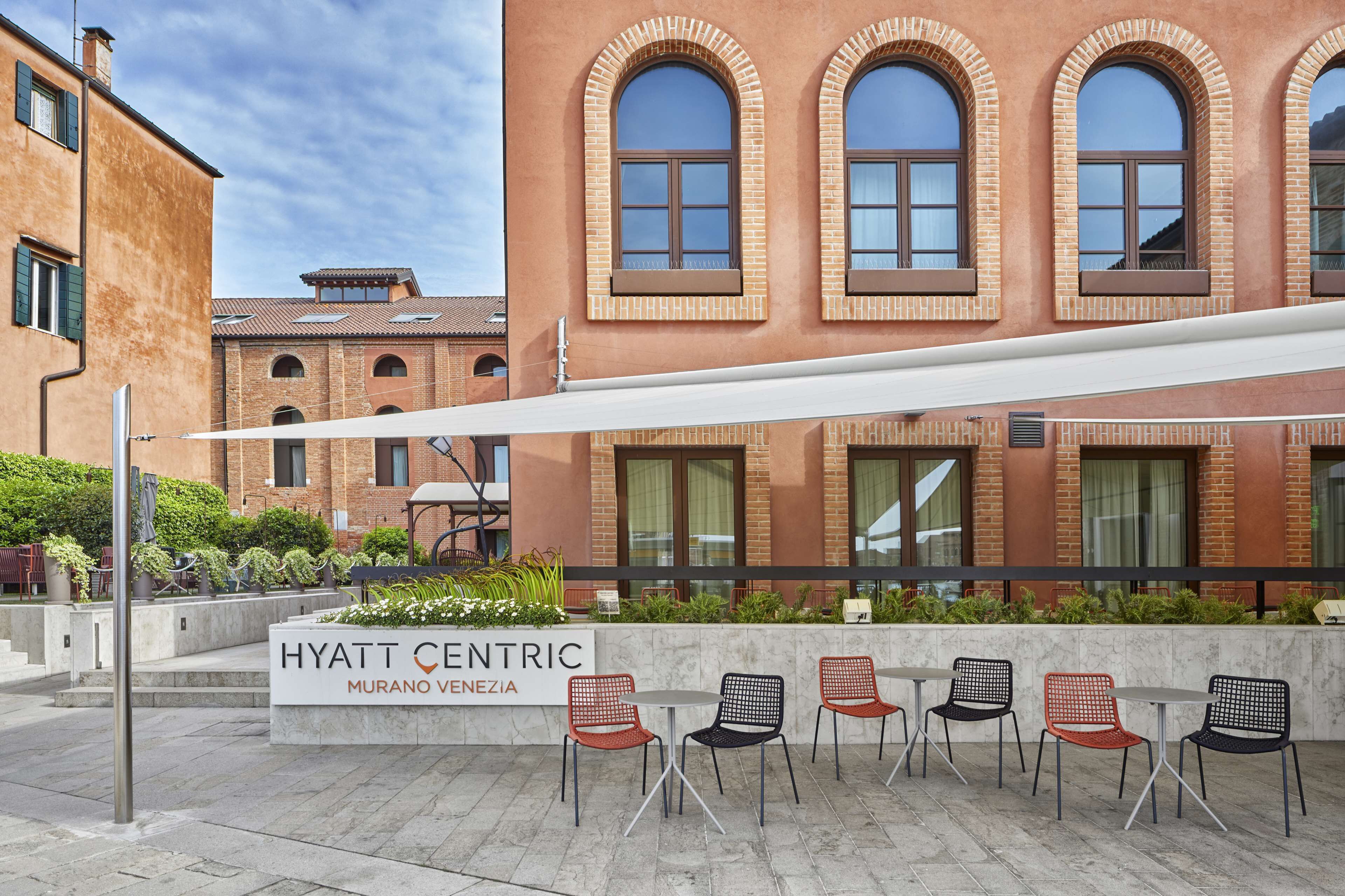 Ein Hotelgebäude mit der Aufschrift 'HYATT CENTRIC MURANO VENEZIA', Sitzgelegenheiten im Freien mit Stühlen und Tischen sowie ein Ziegelhaus mit Rundbogenfenstern.