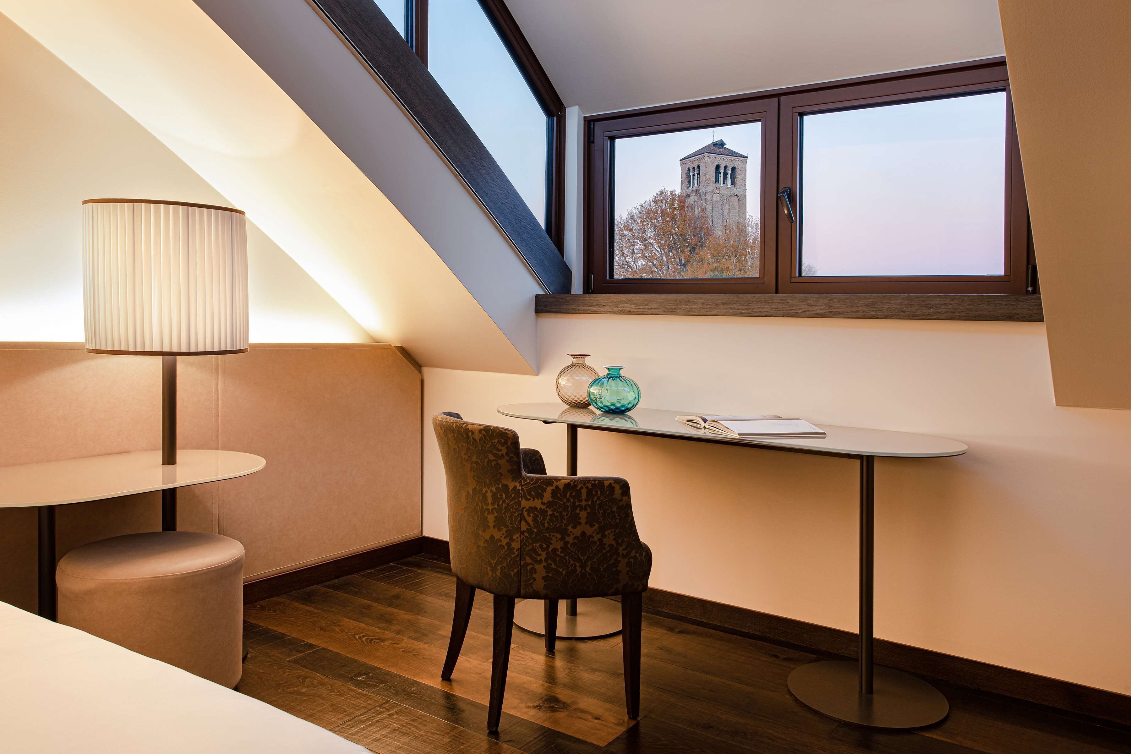 A modern room with a sloped ceiling, a table with a lamp, a chair, and a view of a building through a window.