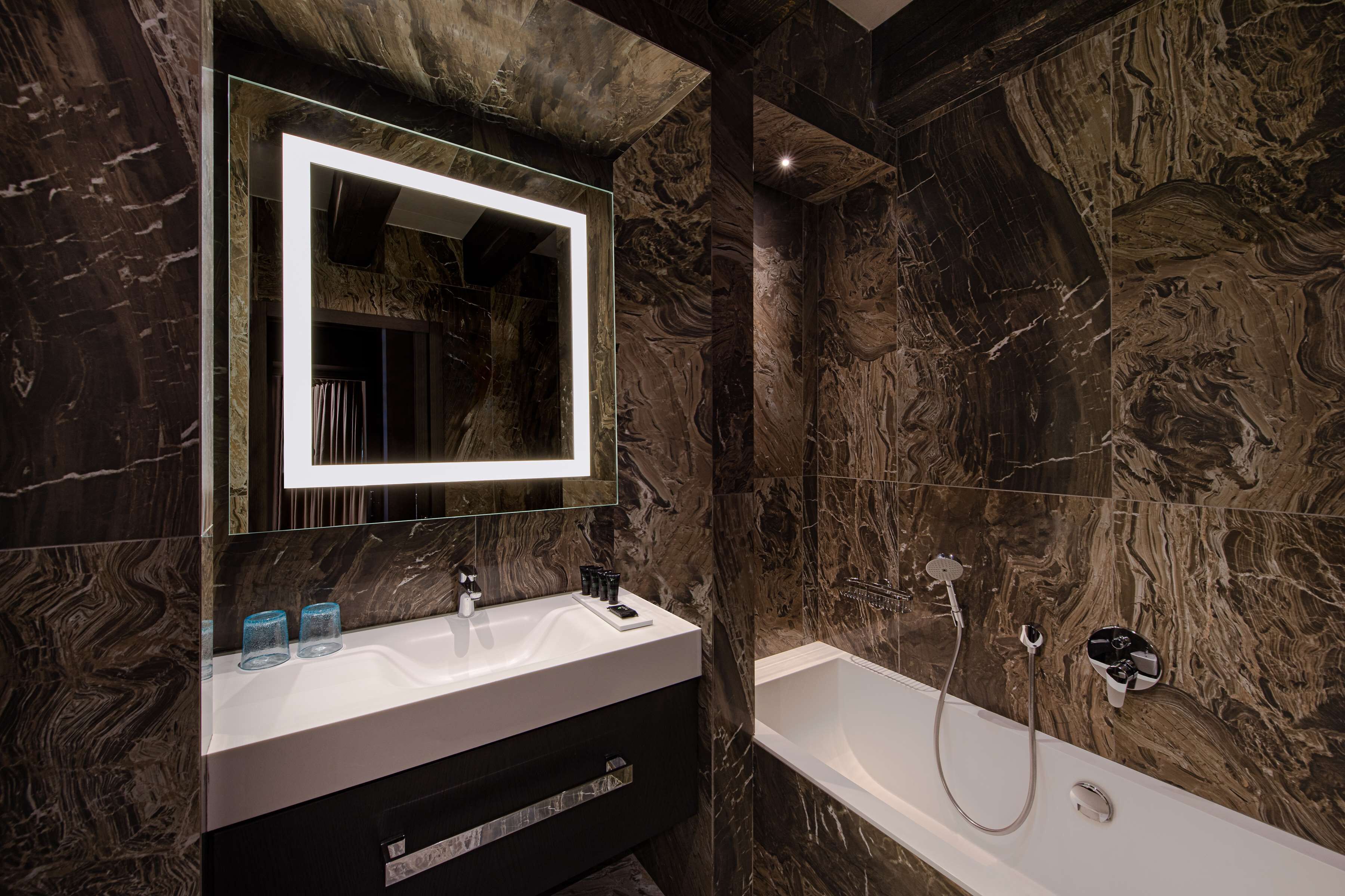 A modern bathroom with a marble wall, a large mirror, a white sink, and a bathtub.