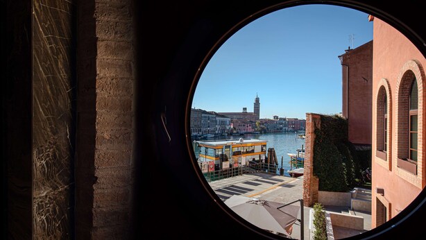 Vista a través de un ventanal circular de una zona ribereña con edificios, un barco y una torre.