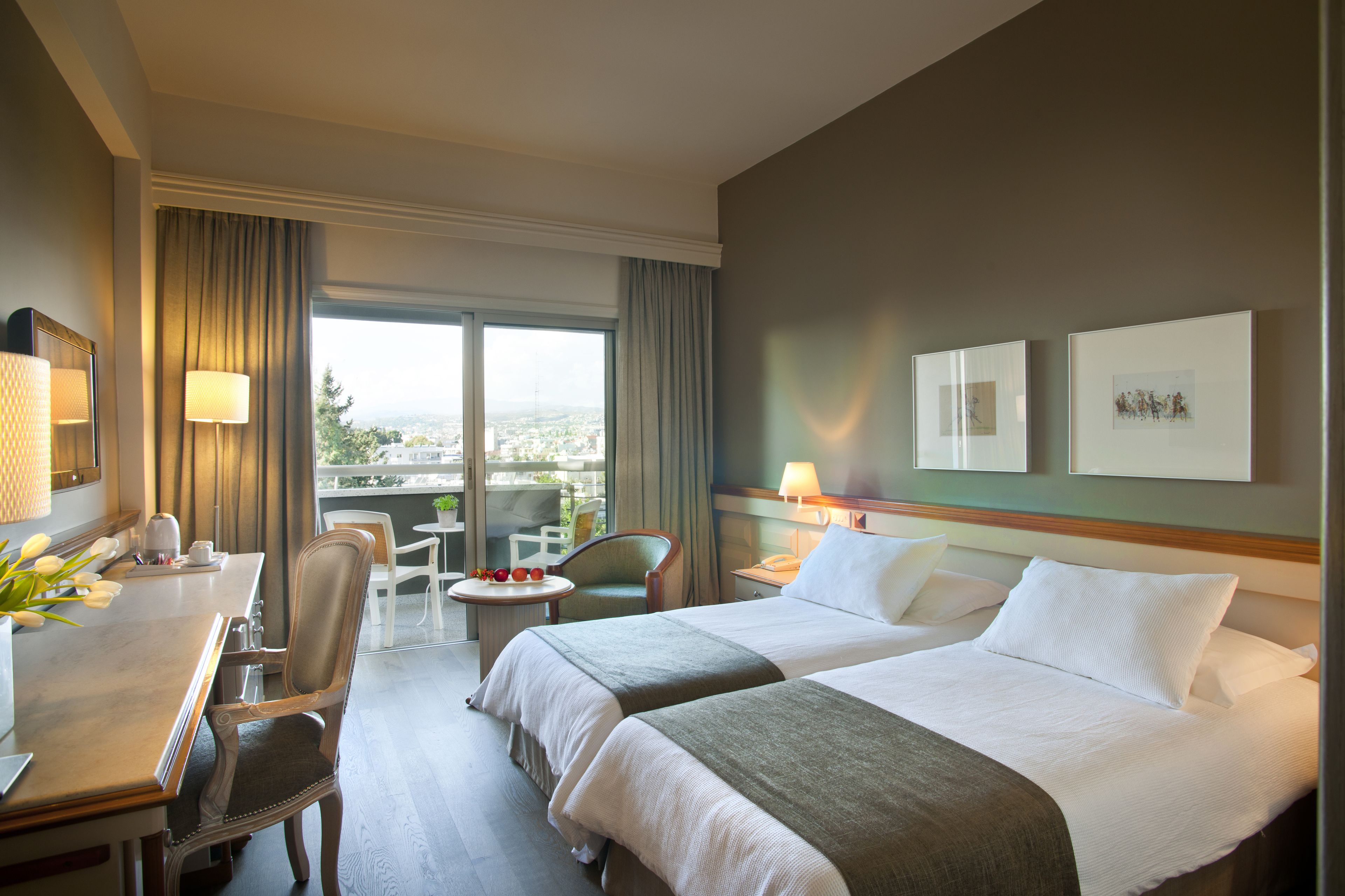 A hotel room with two beds, a desk, a chair, a small table with fruits, and a view of the city.