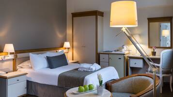 Quarto de hotel com cama, mesas de cabeceira, escrivaninha, cadeira, abajur, espelho e uma pequena mesa com frutas e um vaso.