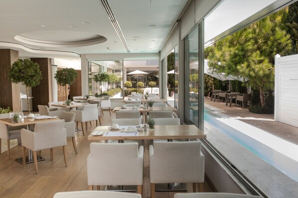 A modern restaurant with indoor seating, large windows, and a view of the outdoor patio area. - Alasia Hotel (Limassol)