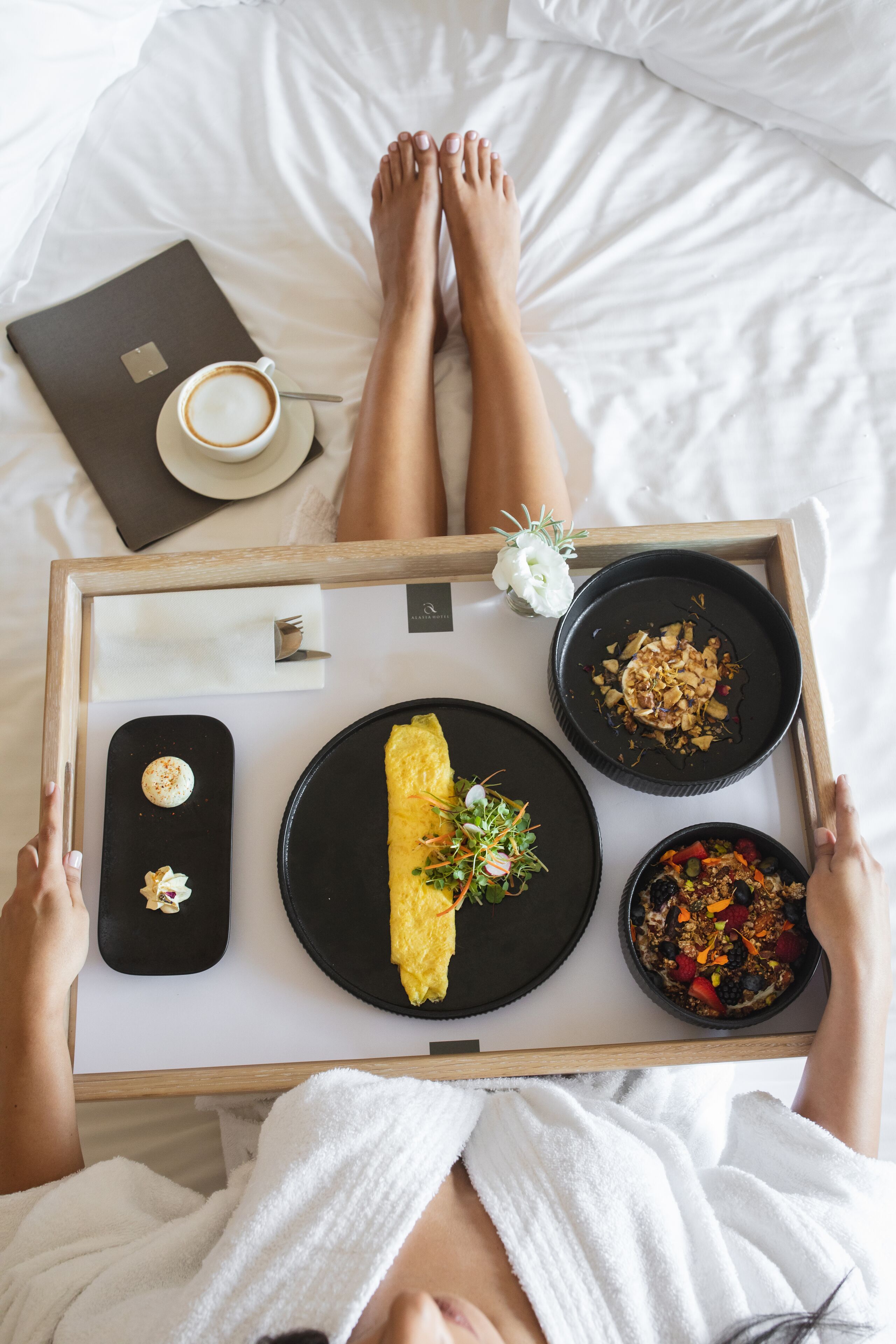 A person lying in bed with a tray of breakfast items including an omelette, a bowl of fruit, and a cup of coffee.