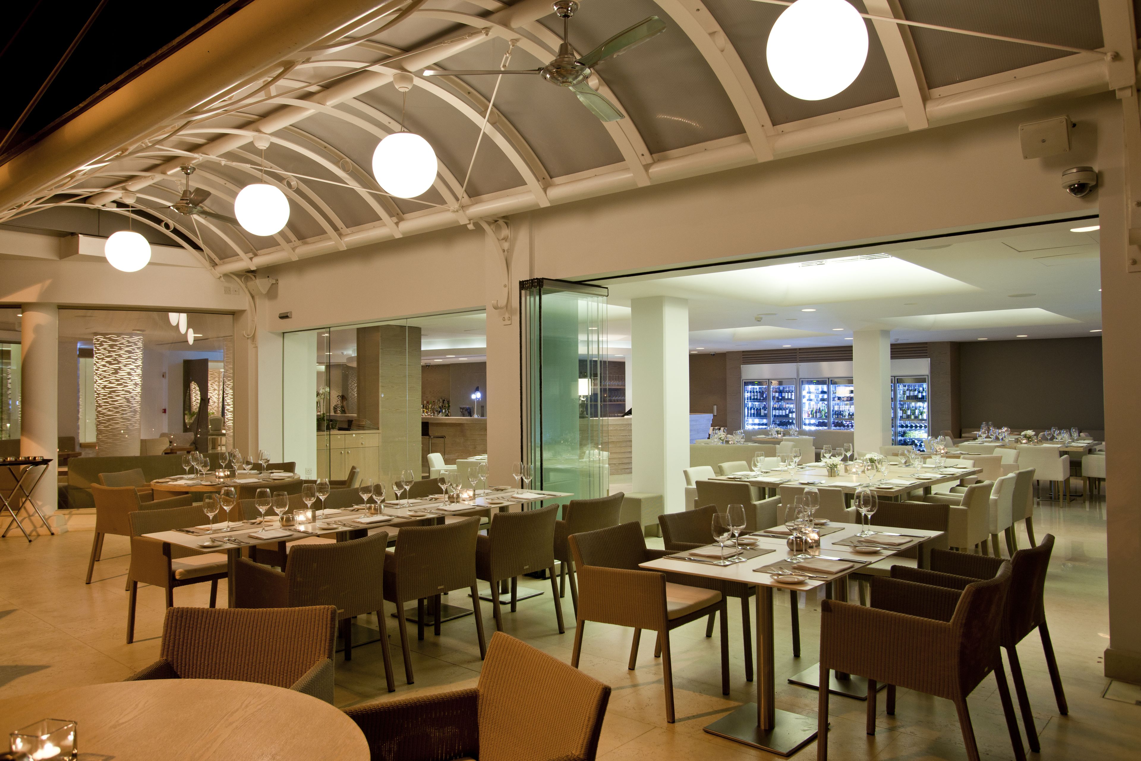 Un restaurante espacioso con varias mesas y sillas, un separador de vidrio que divide el área de comedor de la cocina, y un techo con ventilador e iluminación.