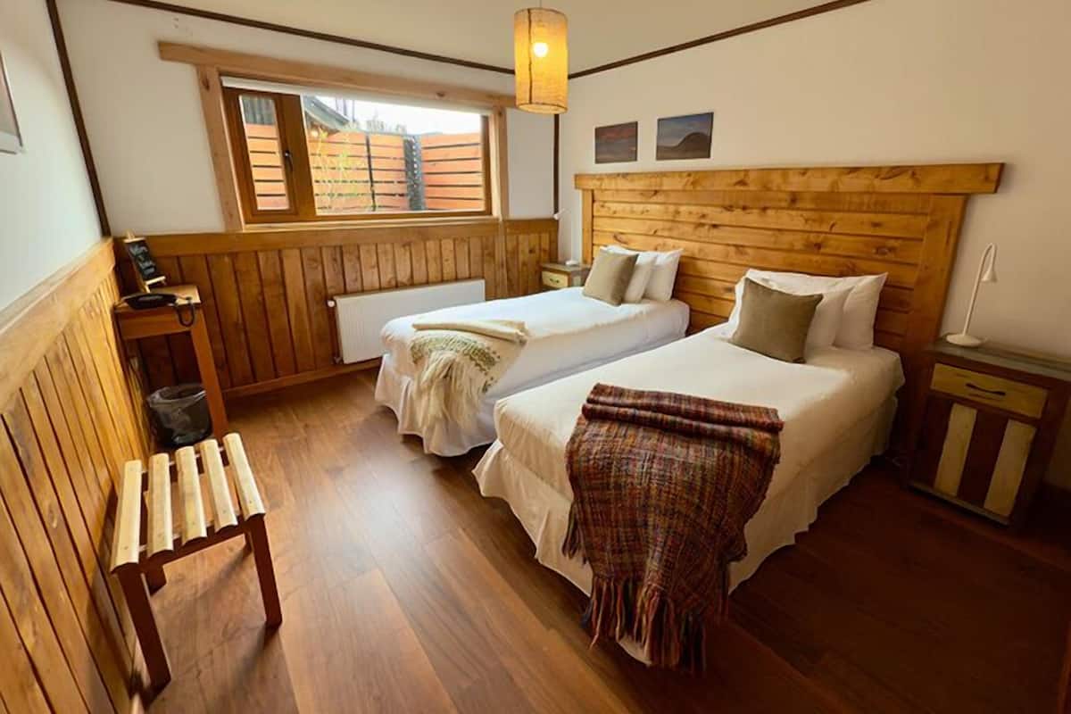 A room with two beds, a wooden bench, a small table, and a window with a view of a fence.