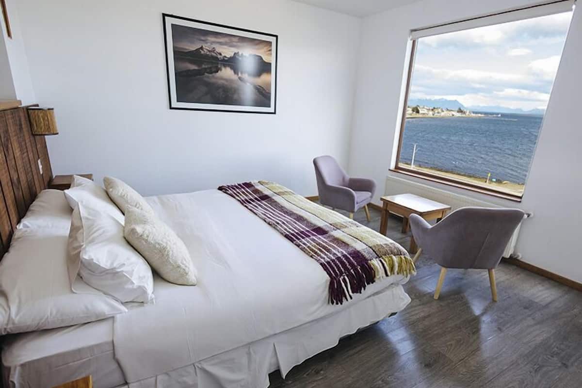 A bedroom with a bed, a chair, a small table, and a large window with a view of the sea.