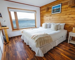 Un dormitorio con un ventanal grande, paredes de madera, una cama con ropa blanca y un cuadro en la pared.