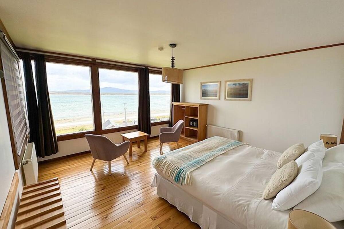 A bedroom with a bed, a desk, a chair, a bookshelf, and a view of the sea.