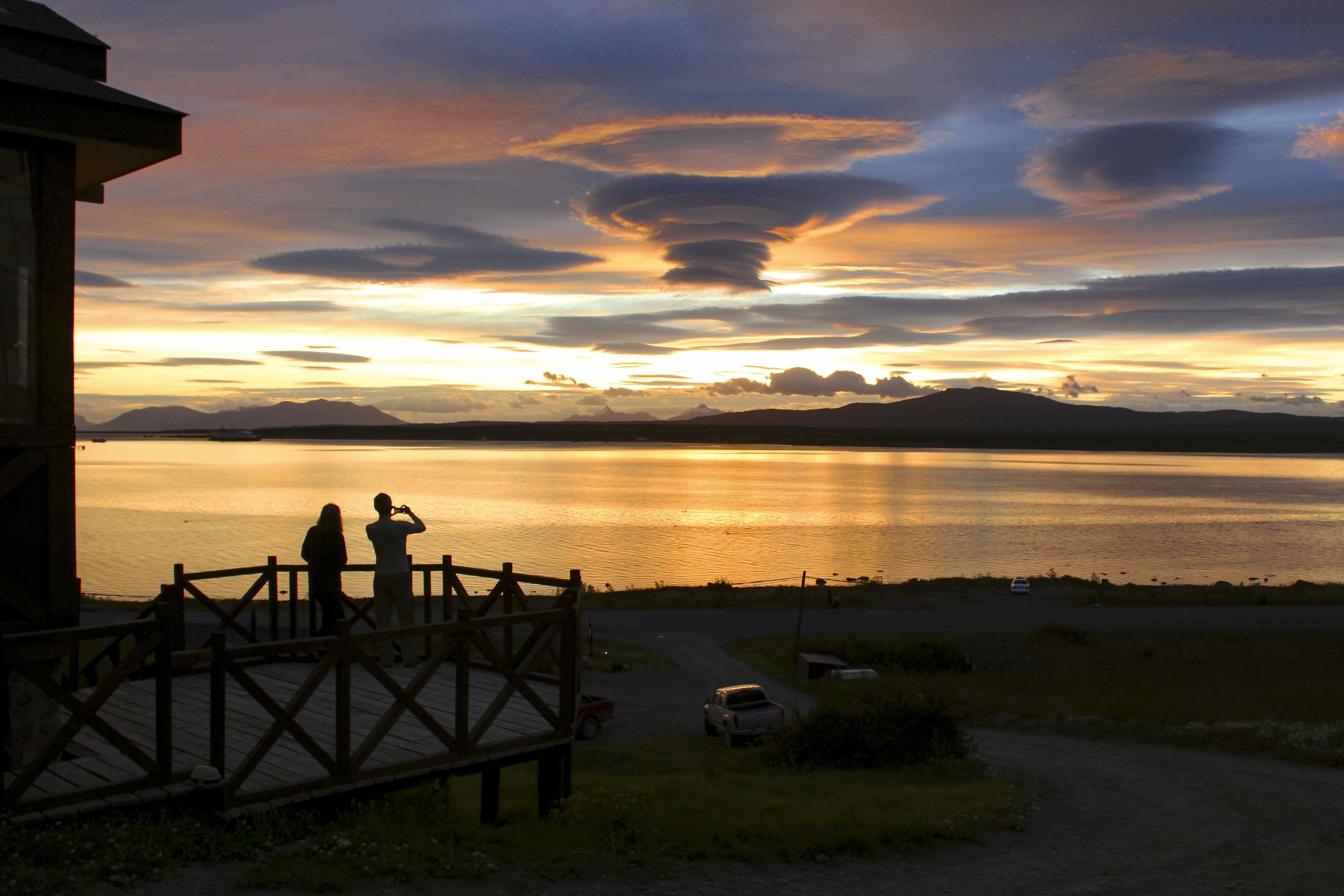 Una vista del atardecer desde una terraza de madera con vista a un lago tranquilo y montañas a lo lejos.
