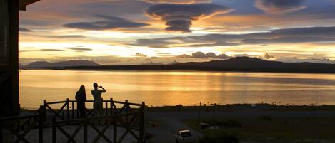 Una vista del atardecer desde una terraza de madera con vista a un lago tranquilo y montañas a lo lejos.