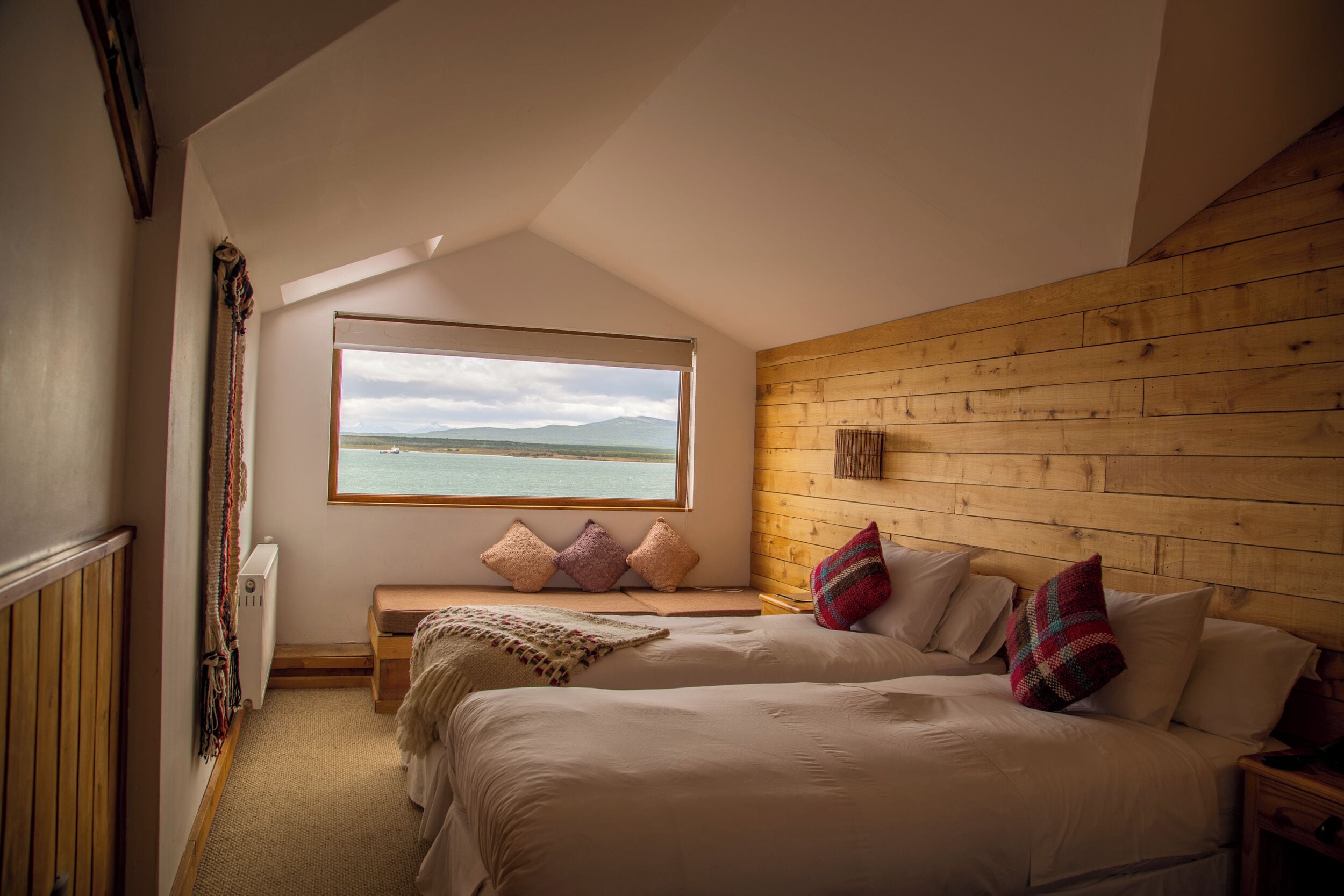 A bedroom with a large window, wooden walls, a bed, a bench, and a view of the sea.