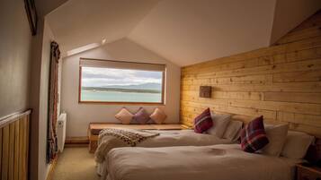A bedroom with a large window, wooden walls, a bed, a bench, and a view of the sea.