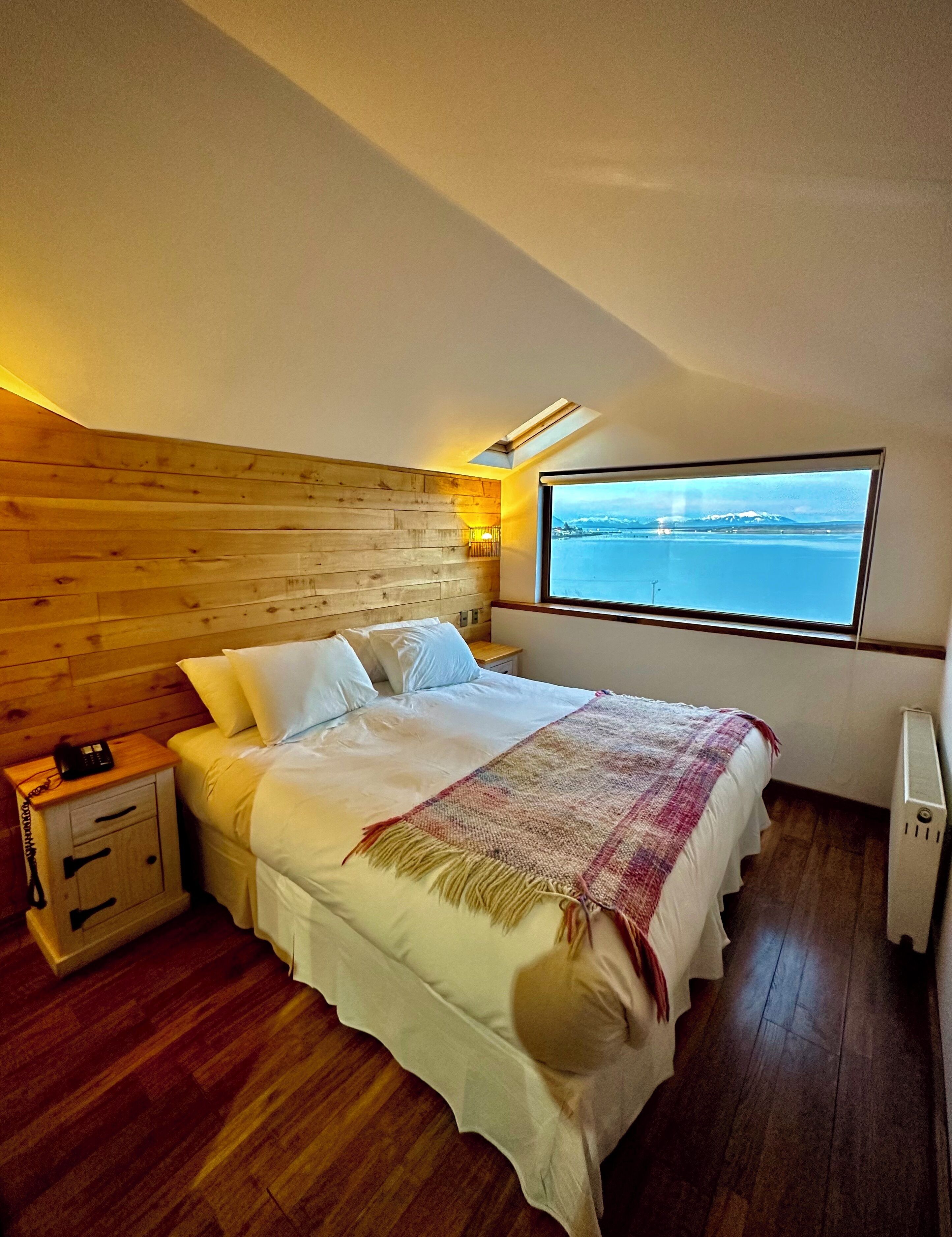 A bedroom with a wooden headboard, a large bed with white bedding, a bedside table with a lamp, and a window with a view of a body of water.