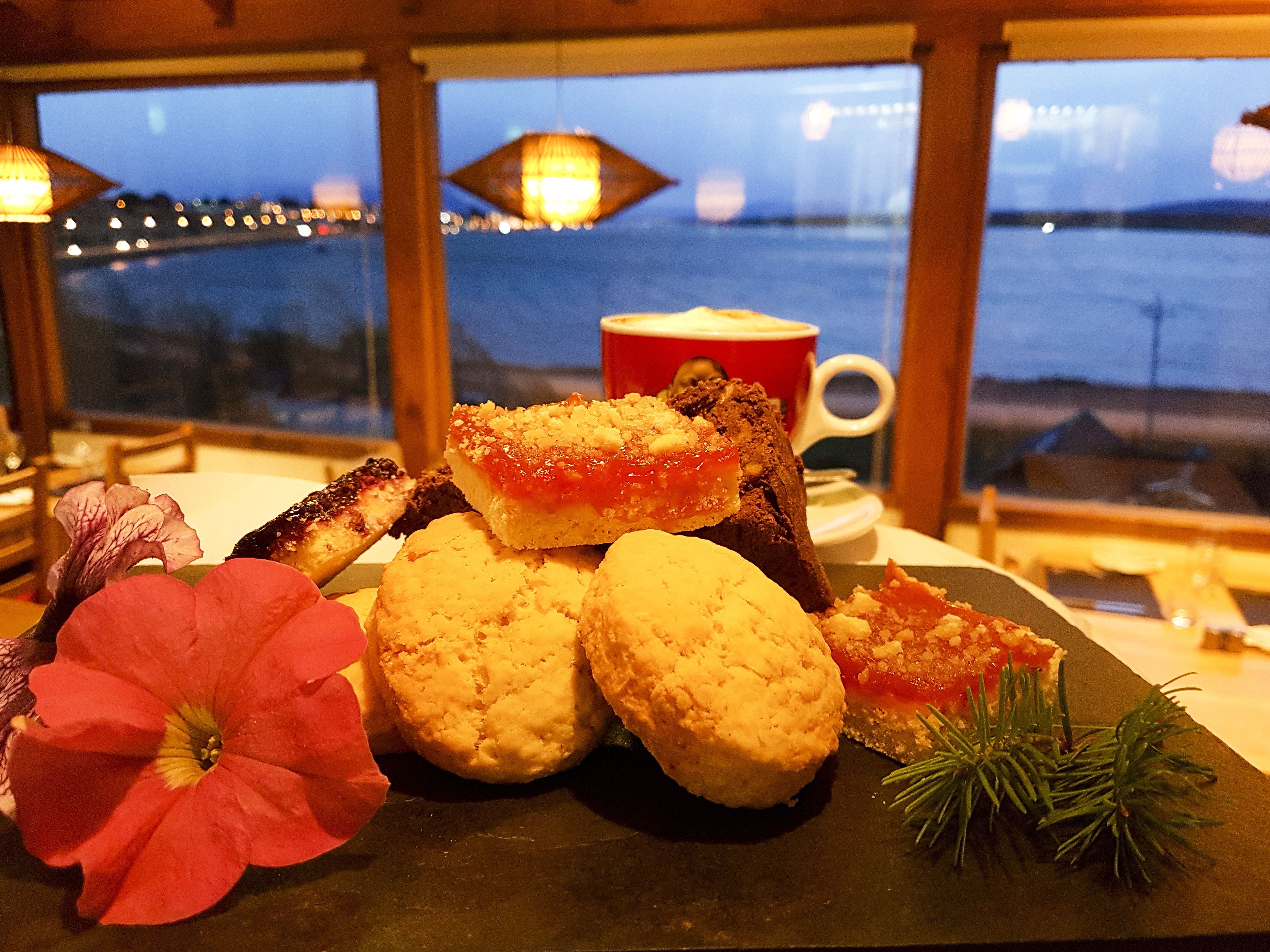 A dessert plate with pastries and a flower, a cup, and a view of a bridge and water at dusk.