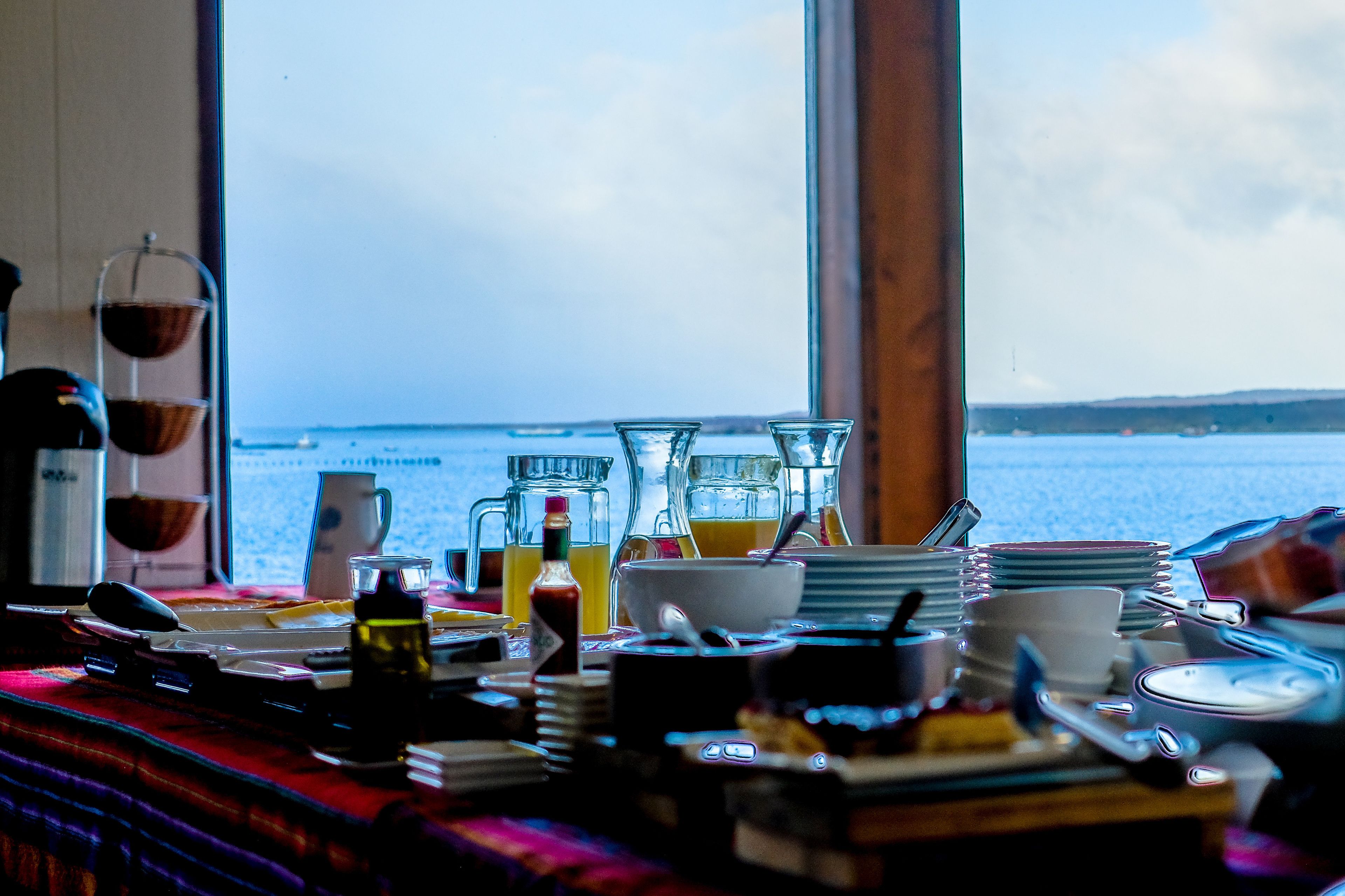 A table with various dishes and drinks, a coffee maker, and a view of the sea through large windows.