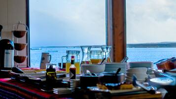 A table with various dishes and drinks, a coffee maker, and a view of the sea through large windows.