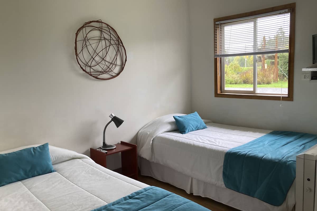 A bedroom with two single beds, a bedside table with a lamp, a window with blinds, and a wall decoration.