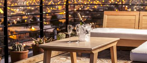 Terraza en la azotea con mesa de madera, dos copas y una botella de champán, con vistas a un paisaje urbano nocturno.