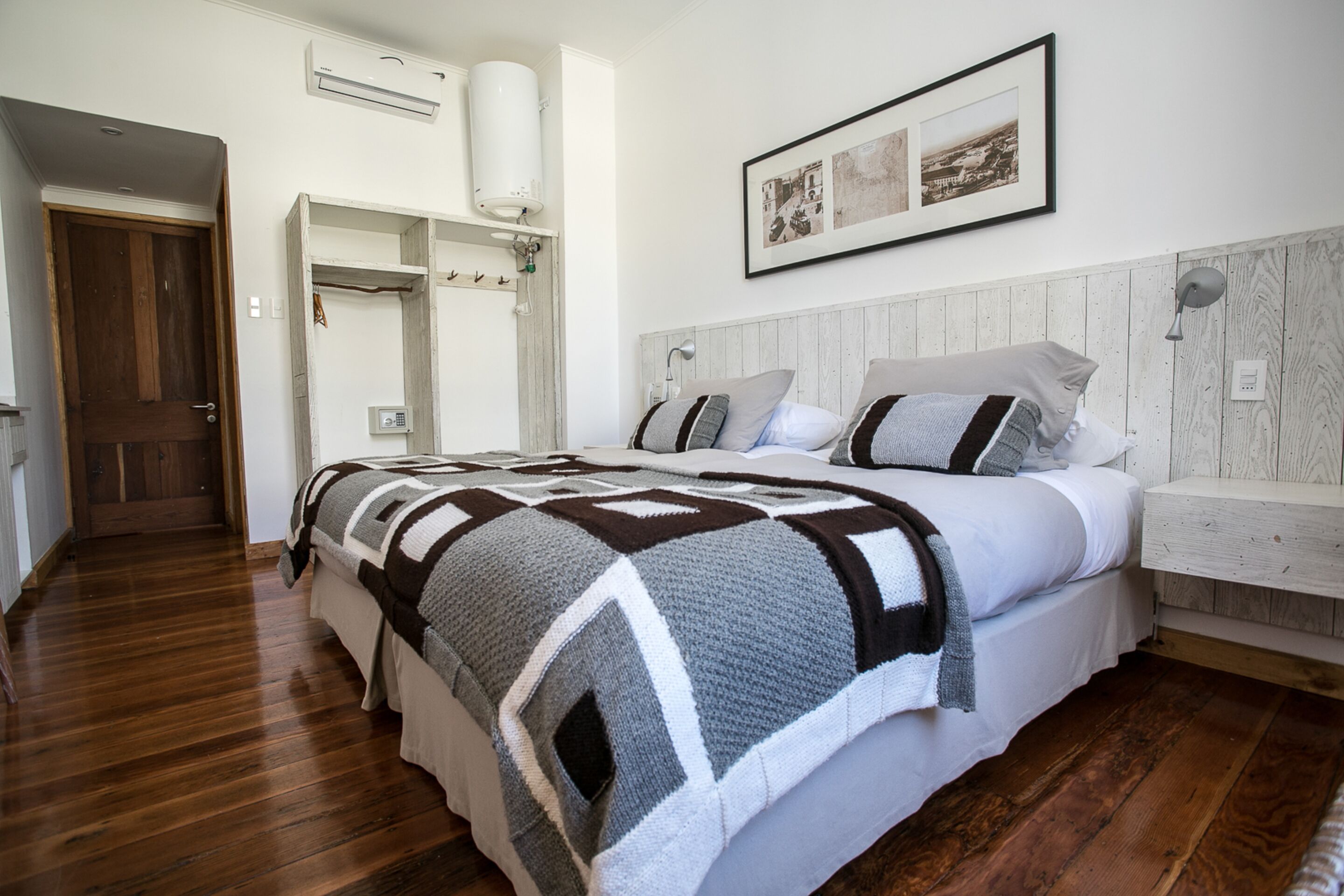 A bedroom with a wooden floor, a bed with a patterned blanket, a white headboard, and a framed picture on the wall.