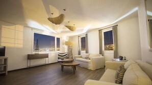 A modern living room with a sofa, armchairs, a coffee table, and a TV. There are large windows with curtains, a lamp, and a view of a cityscape at night. - Casa Galos Hotel & Lofts (Valparaiso)