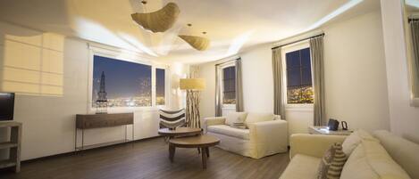 A modern living room with a sofa, armchairs, a coffee table, and a TV. There are large windows with curtains, a lamp, and a view of a cityscape at night.