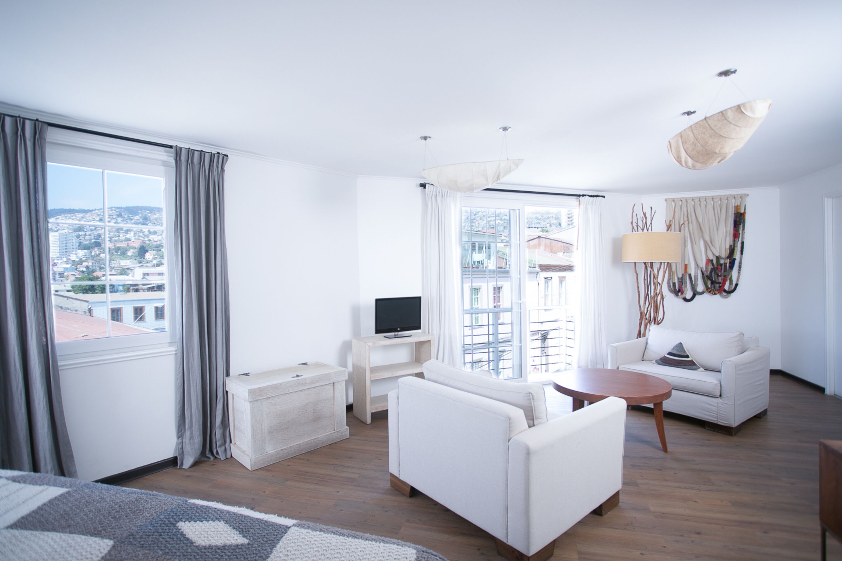 Una habitación de hotel moderna con un ventanal grande, un televisor de pantalla plana, un sofá, una mesa de centro y un tapiz decorativo.