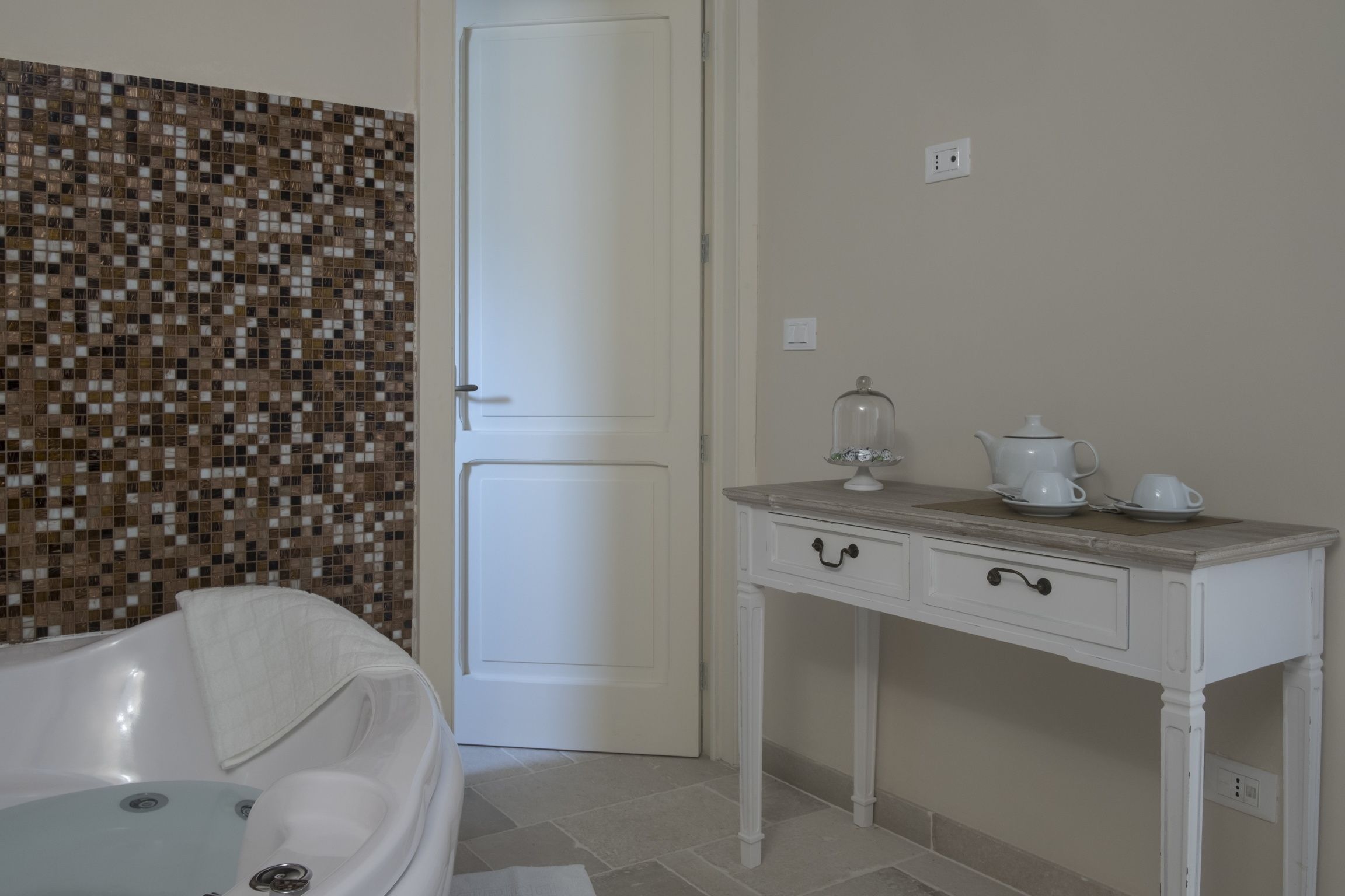 Een badkamer met een wand van mozaïektegels, een witte badkuip, een witte wastafel met spiegel en een witte deur.