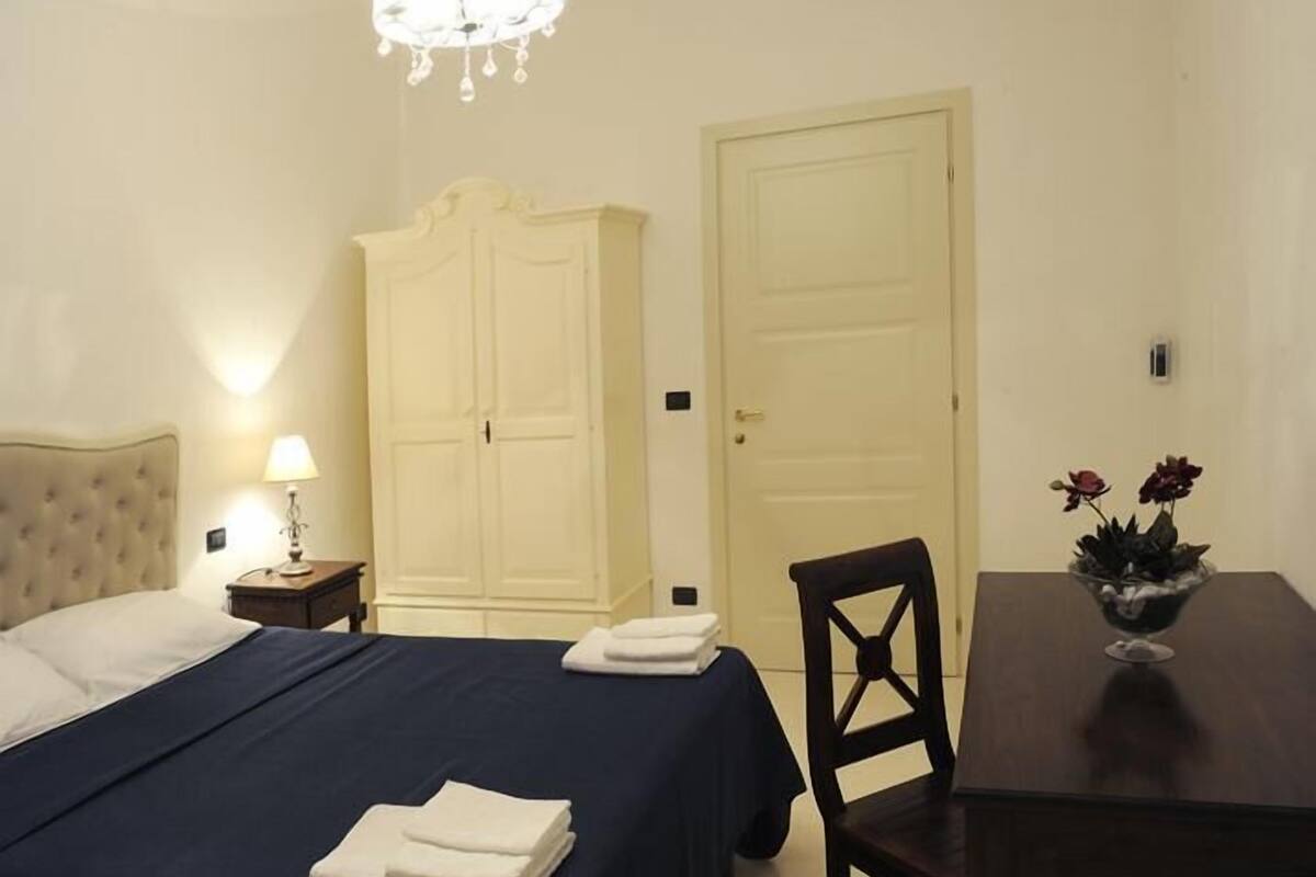 A hotel room with a bed, a nightstand with a lamp, a wardrobe, a dining table with a vase of flowers, and a door.