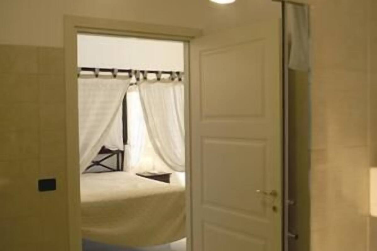 A bathroom with a mirror, sink, and shower. There is a door leading to a bedroom with a bed and curtains.