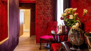 A room with red floral wallpaper, a wooden floor, a red upholstered chair, a small round table with a vase of flowers and two glasses, and a door leading to another room. - Ca Maria Callas (Venice)