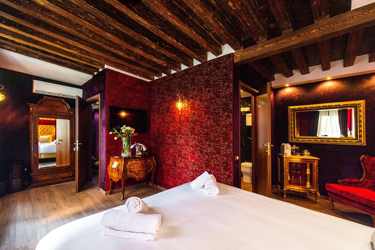 A hotel room with a large bed, a red accent wall, a mirror, and a red chair.