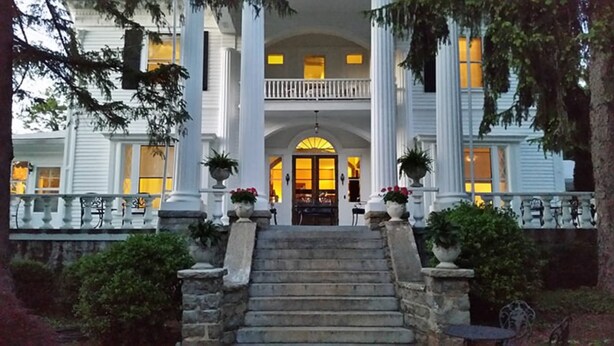 A grand entrance with a staircase leading to a balcony, flanked by columns and potted plants.