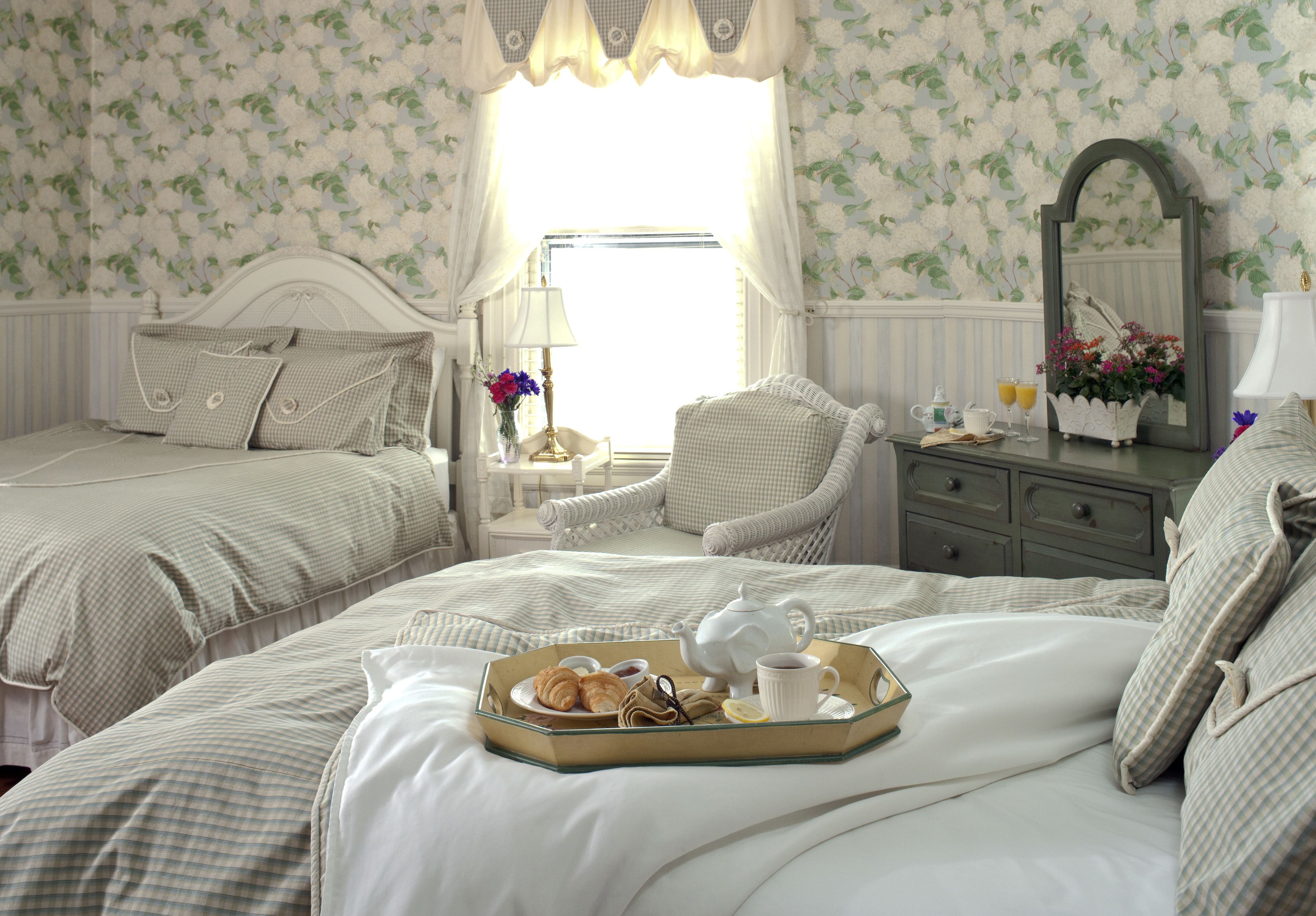 A neatly arranged bedroom with two beds, a bedside table with a lamp, a chair, and a tray with breakfast items.