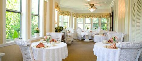 A room with white wicker chairs and tables, large windows, and ceiling fans.
