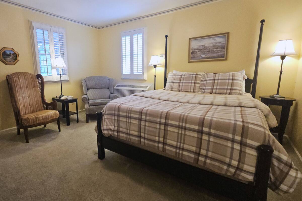 A bedroom with a bed, two chairs, a desk, a lamp, and a painting on the wall.