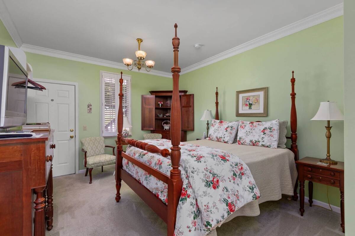 A bedroom with a four-poster bed, a wooden dresser, a chair, a bedside table with a lamp, and a window with blinds.