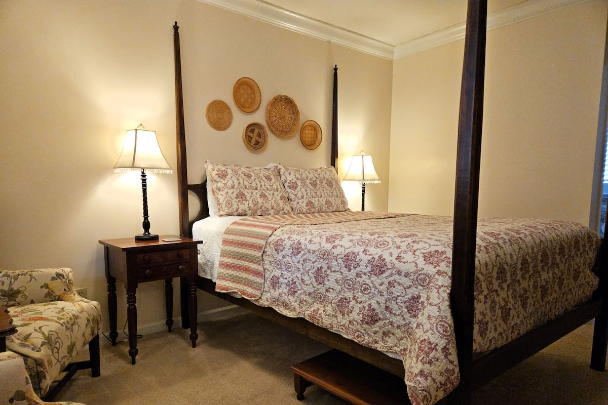 A four-poster bed with a floral bedspread, two bedside tables with lamps, a small chair, and a wall with decorative plates.