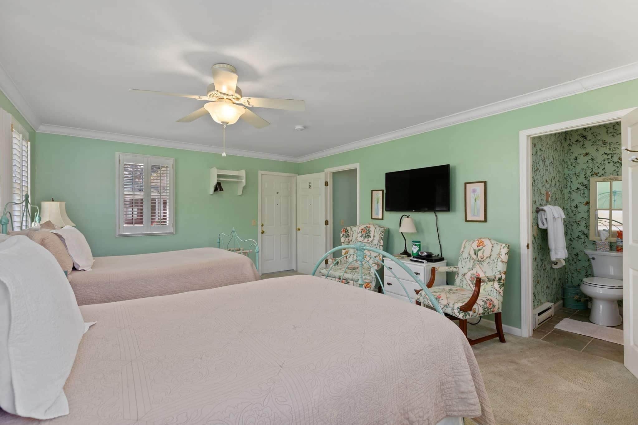 A bedroom with two beds, a ceiling fan, a television, and a bathroom with a toilet and sink.