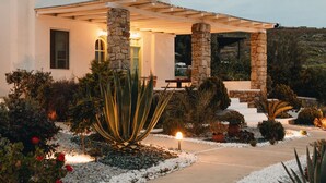 A modern house with a stone facade, a covered patio, and a landscaped garden with various plants and a pathway. - Mykonos Pantheon (Mykonos)