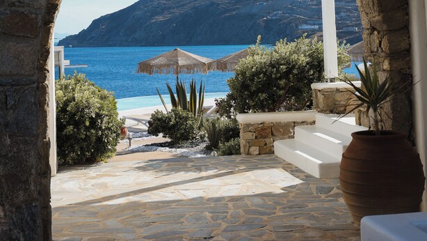 An arched stone doorway leading to a stone-paved area with a view of a blue sea and a mountainous landscape. - Mykonos Pantheon (Mykonos)