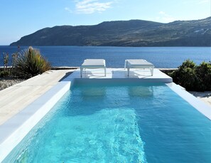 A swimming pool with two white lounge chairs, surrounded by a wooden deck, with a view of the sea and hills in the background.