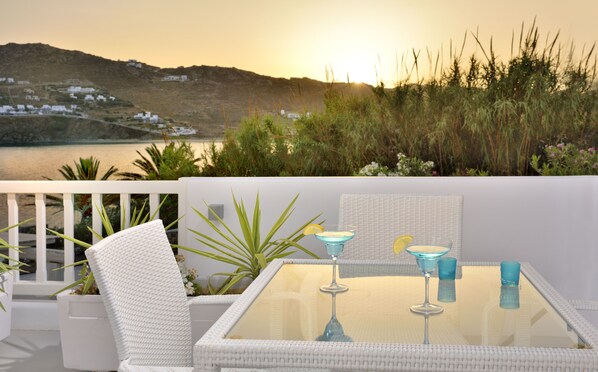 Un balcón con una mesa de vidrio, sillas de mimbre blanco y vistas a un paisaje costero con edificios y vegetación.
