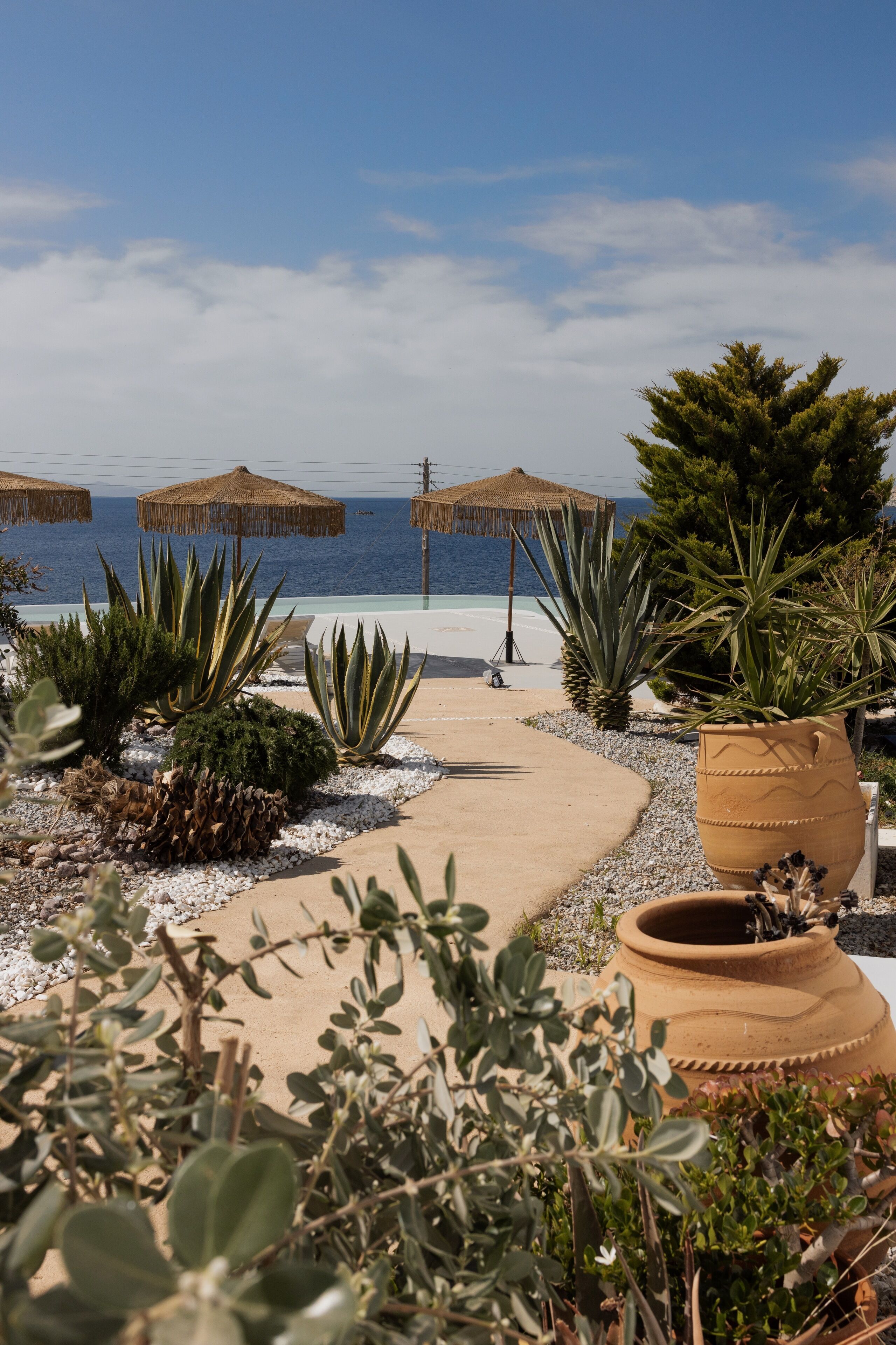 A pathway leading to a beach with thatched umbrellas, potted plants, and a view of the sea.