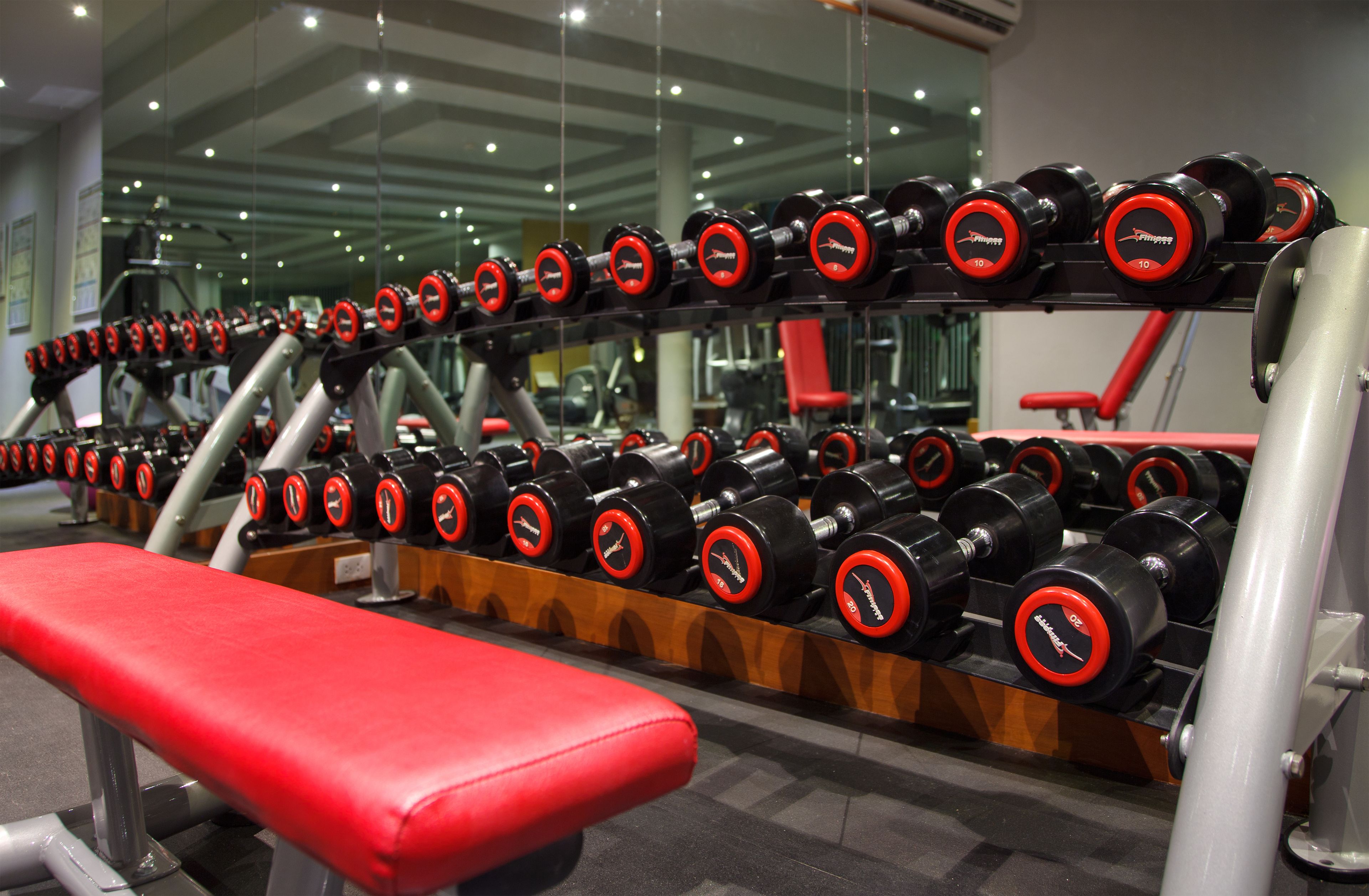 A gym with rows of red and black dumbbells on racks, a red bench, and various exercise machines.
